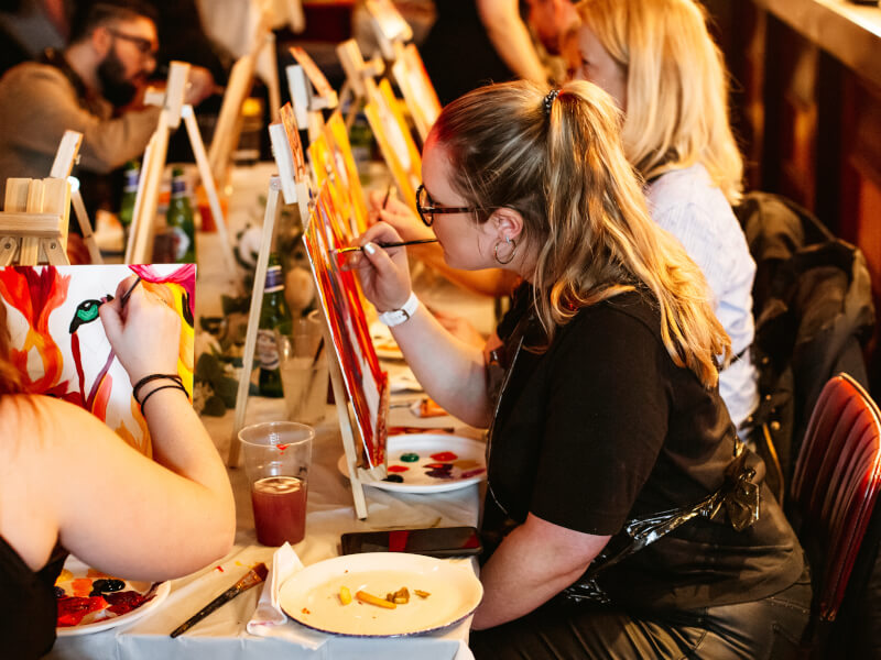 People sat painting on table top canvas and easels at a colourful art class.